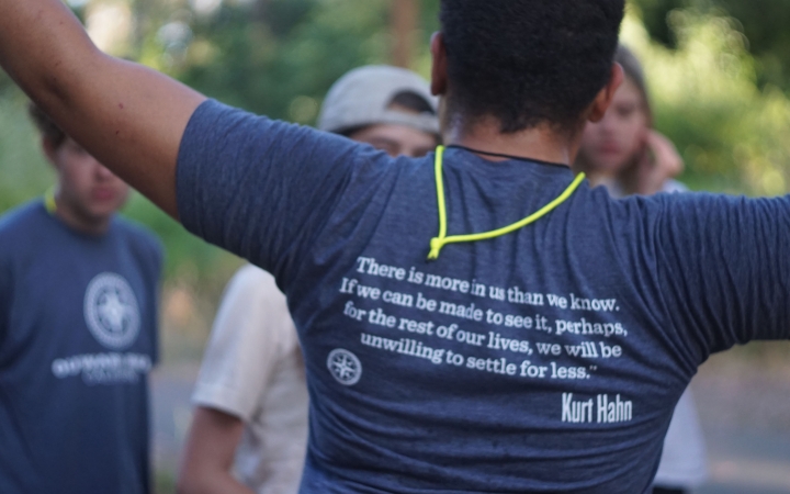 A person wearing an outward bound shirt lifts their hands in the air. The back of the shirt reads, "There is more in us than we know. If we can be made to see it, perhaps for the rest of our lives, we will be unwilling to settle for less. – Kurt Hahn"